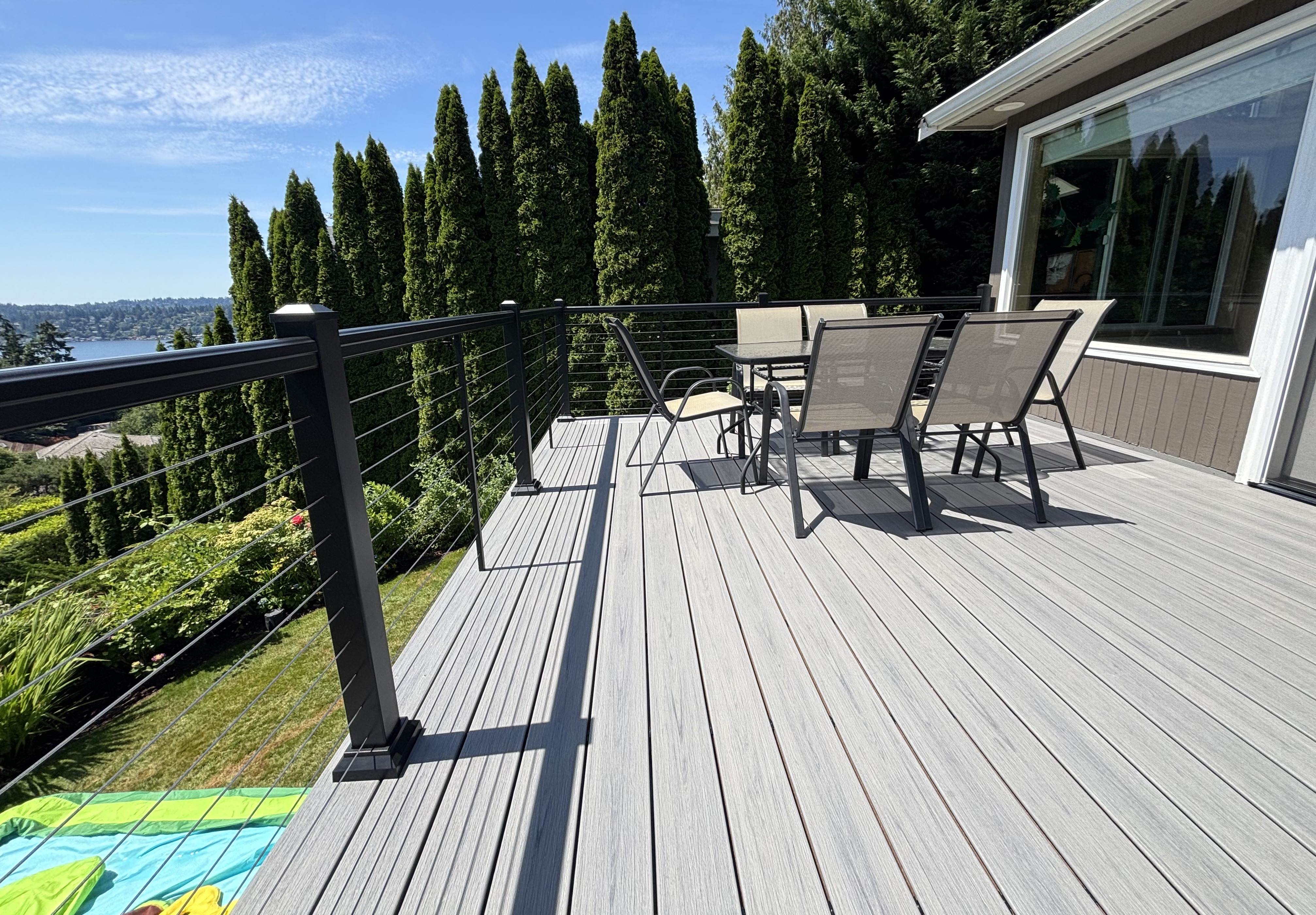 Finished light-gray composite deck with sleek black cable railing and an outdoor dining set, overlooking a landscaped garden and lake view on a sunny day